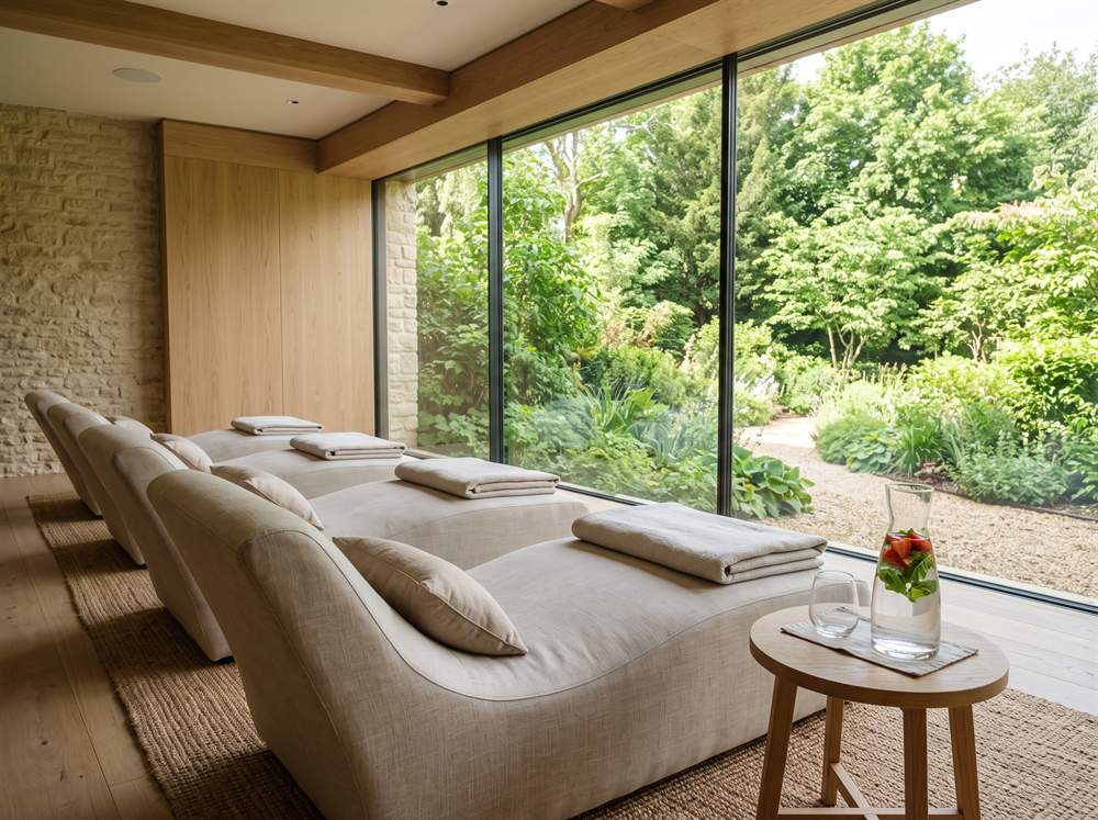 Ruheraum eines Hotel-Wellnessbereichs mit Liegen am Panoramafenster zum Garten, auf dem Tischchen eine Karaffe Wasser mit Erdbeeren und Minze